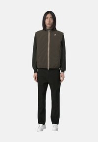 Gilet imbottito marrone con colletto alto, dotato di zip frontale e dettagli a strisce multicolori, indossato sopra una camicia verde a maniche lunghe e pantaloni neri.