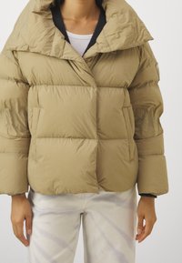 Beige pufferväst med bred krage, stora vadderade partier och sidofickor. Materialet är slätt och lätt glänsande.