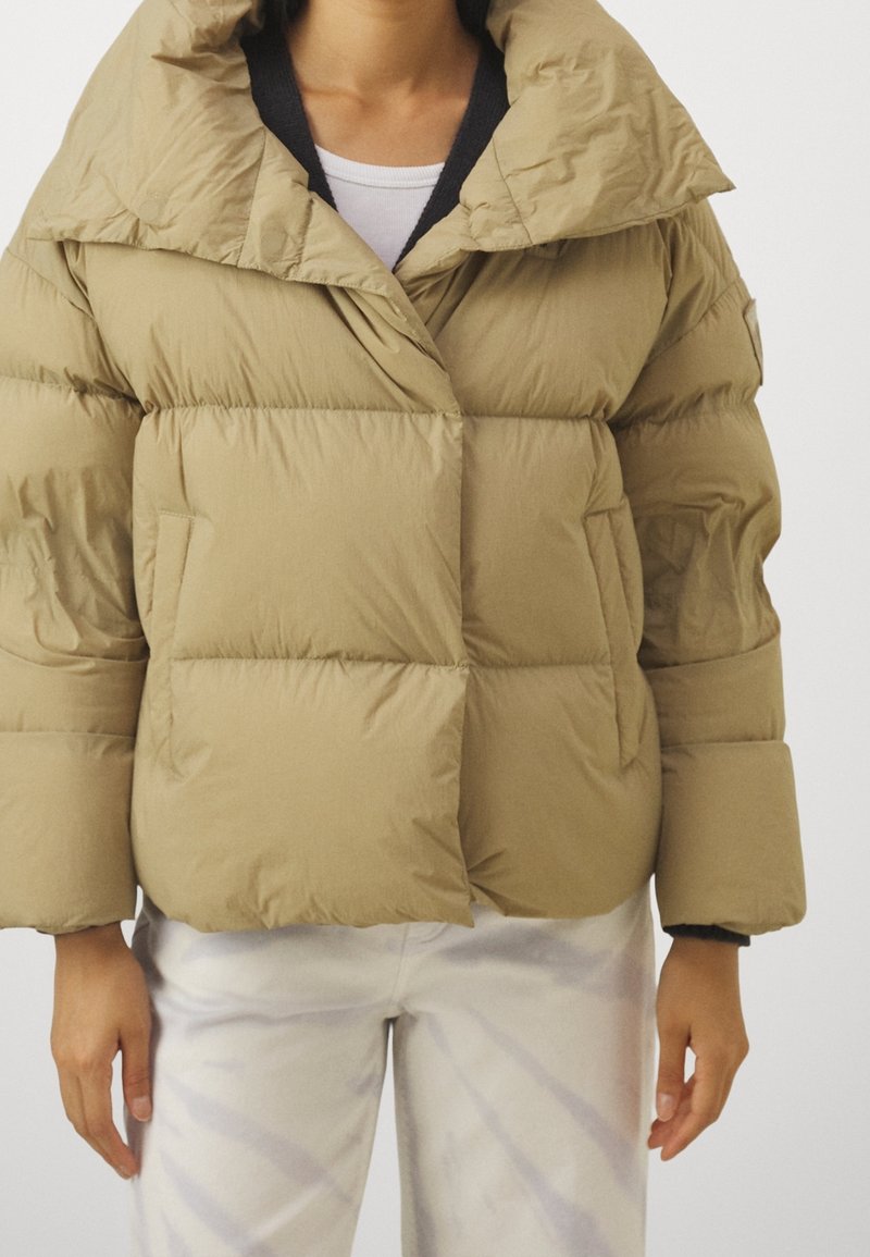 Beige pufferväst med bred krage, stora vadderade partier och sidofickor. Materialet är slätt och lätt glänsande.