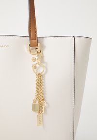 Sac fourre-tout en matière texturée crème avec une poignée marron, agrémenté de quincaillerie dorée et d'une chaîne avec des charms de cadenas et de clé.