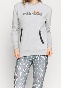 Sudadera gris con bolsillos con cremallera negros, que presenta el logo de "ellesse" y un gráfico de colores encima. Combinada con mallas grises con patrones en blanco y burdeos.
