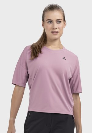 Frau mit hellbraunem Pferdeschwanz trägt ein locker sitzendes, lavendelfarbenes Kurzarmshirt und schwarze Hose, blickt leicht zur Seite.