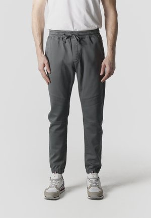 Hombre con pantalones jogger gris oscuro con puños elásticos y cintura con cordón, zapatillas blancas y camiseta blanca, de pie frente a un fondo neutro.