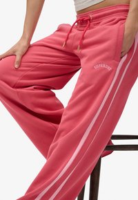 Donna che indossa pantaloni della tuta Superdry rosa con strisce laterali rosa chiaro, appoggiando una mano sul ginocchio e l'altra in tasca mentre è seduta.
