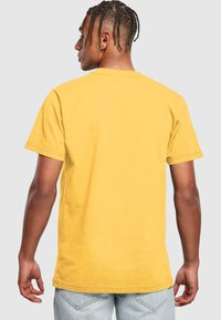 Geltona medvilnė t-shirt su apykakle ir trumpomis rankovėmis, rodoma iš nugaros, pasižyminti laisvu siluetu ir tiesiu kraštu.