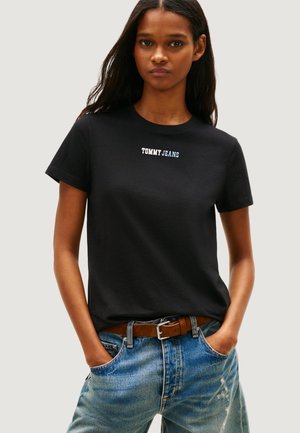 Žena v černém tričku Tommy Jeans zastrčeném do modrých džínů s hnědým opaskem, ruce v kapsách, neutrální pozadí.