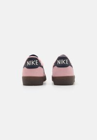 Nike sportcipők rózsaszín velúrból készült felsőrésszel és sötétkék részletekkel. Jellemzője egy textúrált barna talp és a kiemelkedő "NIKE" márkázás a saroknál.