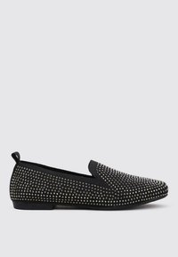 Zapatos negros tipo slip-on cubiertos de detalles metálicos, con una parte superior de tela suave y una suela plana de goma. Sin costuras visibles ni adornos.