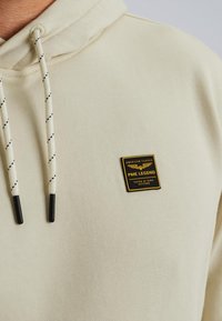 Bež dukserica s crno-bijelim vezicama i malim crno-žutim logom koji glasi "PME Legend, American Classic, Tested at High Altitude."
