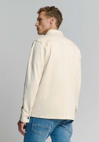 Crème-kleurige jas met een kraag, zichtbare naden en knopenmanchetten. Draagt bij blauwe spijkerbroeken. Soepele stoftextuur.