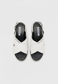 Hvide sandaler med krydsrem og sort sål, med spænde lukning og "Karl Lagerfeld" branding på remmen. Glat tekstur.