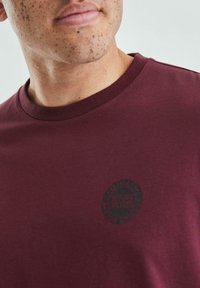 Bordowa, krótko-sleeve koszulka T-shirt wykonana z bawełny, z czarnym okrągłym logo na klatce piersiowej oraz okrągłym dekoltem. Miękka, gładka tekstura.