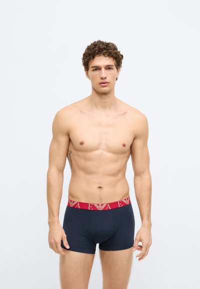 Cuecas boxer azul-marinho com uma cintura elástica vermelha com um logótipo. Tecido suave e elástico; corte ajustado. Modelo está de pé contra um fundo simples.