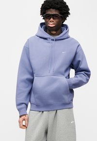 Μωβ φούτερ Nike με μπροστινή τσέπη, κουκούλα με κορδόνι και ριμπ μανσέτες, συνδυασμένο με γκρι φόρμες που διαθέτουν μικρό λογότυπο.