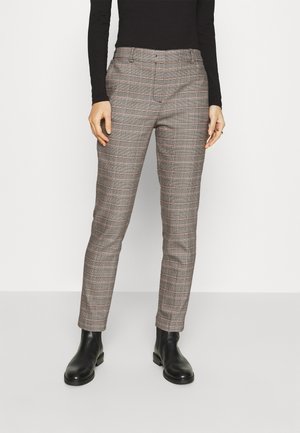 Vrouw die slanke beige en grijs geruite broek draagt, gecombineerd met zwarte enkellaarsjes en een zwart shirt met lange mouwen, staand tegen een effen achtergrond.