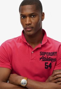 Rød polo skjorte med krave, med mørkeblå broderet tekst "SUPERDRY Athletique 54." Guld ur med læderrem på håndleddet.