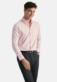 Jasnoróżowa koszula z długim rękawem, z kołnierzykiem zapinanym na guziki, z przodu listwy i haftowanym logiem, zestawiona z ciemnymi spodniami i czarnym paskiem.