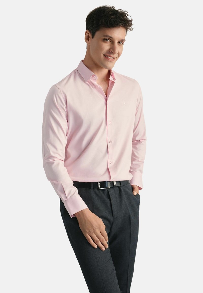 Jasnoróżowa koszula z długim rękawem, z kołnierzykiem zapinanym na guziki, z przodu listwy i haftowanym logiem, zestawiona z ciemnymi spodniami i czarnym paskiem.