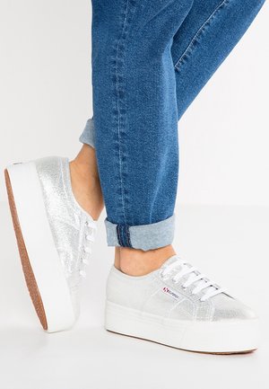 Persona que lleva vaqueros azules remangados y zapatillas plataformas plateadas con cordones blancos y suelas gruesas blancas, de pie sobre suelo blanco.