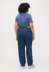 Oseba s kodrastimi lasmi nosi modro majico s kratkimi rokavi, modre hlače z visokim pasom in bele superge, stoji obrnjena stran od preproste ozadja.