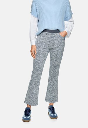 Person, der blaue, strukturierte Hosen, ein blau-weißes, mehrlagiges Oberteil und blaue Turnschuhe trägt und mit einer Hand in der Tasche steht.