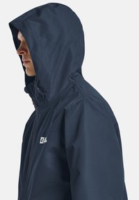 Giacca impermeabile blu navy con cappuccio, dotata di zip frontale e polsini elasticizzati. Texture liscia con un branding discreto sul petto.