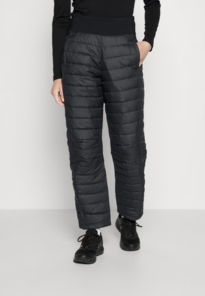 Person trägt schwarze gesteppte, gepolsterte Hose, schwarzen Oberteil, schwarze Turnschuhe, steht mit einer Hand in der Tasche vor einfachem Hintergrund.