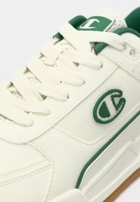 Λευκό αθλητικό παπούτσι με πράσινες λεπτομέρειες, κατασκευασμένο από δέρμα και δίχτυ, με επίπεδα κορδόνια, υφασμένο πάνελ και λογότυπο στο πλάι.