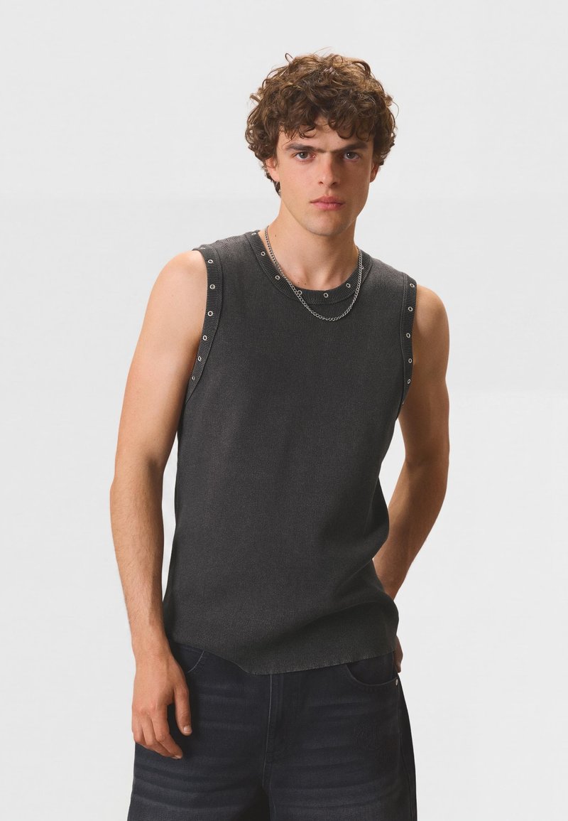 Jeune homme aux cheveux bouclés portant un débardeur gris foncé avec des détails œillets et un collier chaîne en argent, debout devant un fond uni.