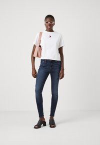 Camiseta blanca corta con un pequeño logo, jeans oscuros ajustados, mules negras de tacón bloque abierto y un bolso de hombro rosa.