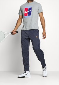 Graues Baumwoll-T-Shirt mit einem blauen und roten Nike-Logo, kombiniert mit marineblauen, strukturierten Hosen und weißen Nike-Schuhen. Hält einen Tennisschläger.