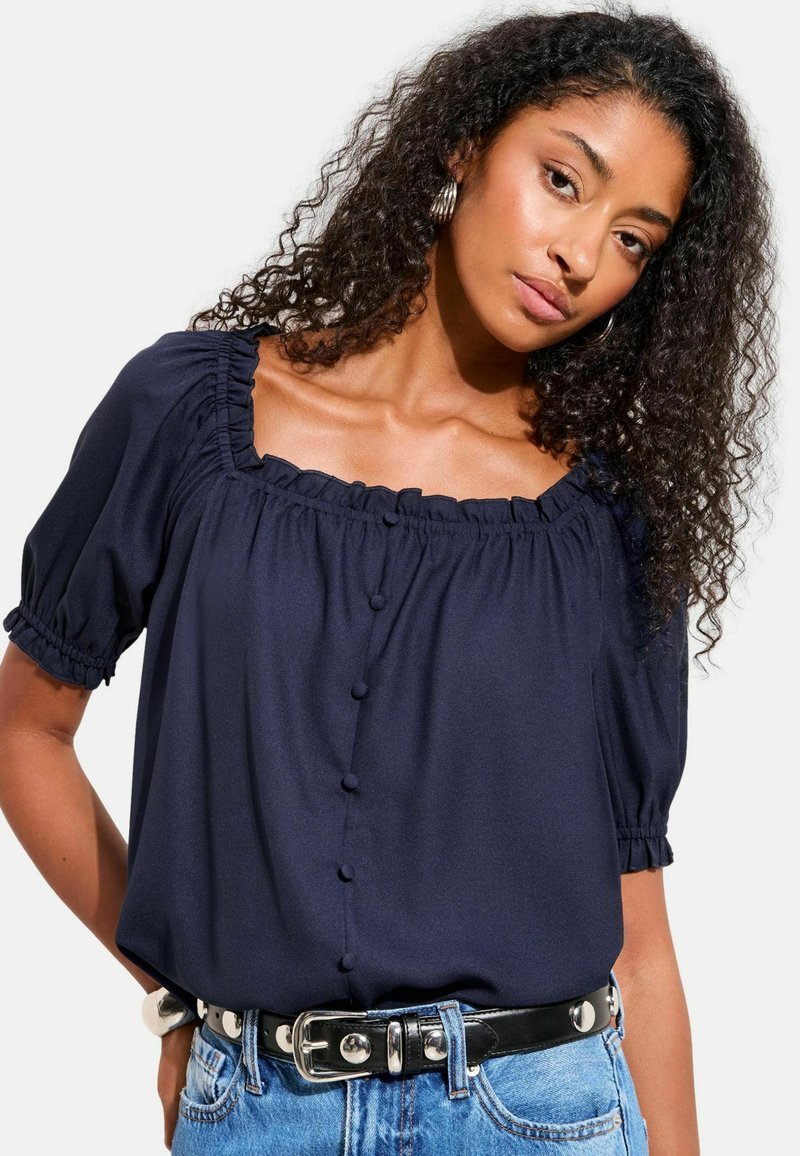 Vrouw met krullend haar, gekleed in een marineblauwe blouse met ruches en een vierkante halslijn met knopen, en een spijkerbroek met een zwarte riem met zilveren studs.