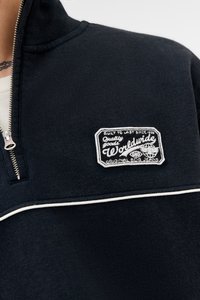 Nærfoto af en mørk zip-up sweater med hvid piping og et patch med teksten "Quality goods Worldwide Original SPG Jeans."