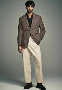 Brauner, strukturierter Blazer über einem schwarzen Hemd, kombiniert mit einer cremefarbenen Hose und dunklen Schuhen. Vor einem neutralen Hintergrund präsentiert.
