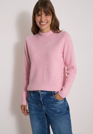 Femme souriante portant un pull côtelé rose clair à manches longues et un jean bleu, une main dans la poche, sur un fond uni.