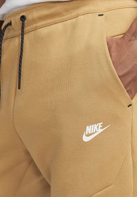 Khaki Nike sweatpants with a smooth texture, featuring black drawstrings, a white logo on the thigh, and side pockets.