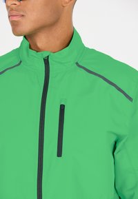 Veste légère verte avec un col haut, fermeture éclair à l'avant et une poche poitrine. Présente des accents gris le long des épaules. Texture lisse.