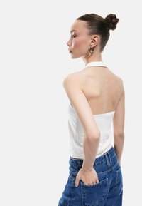 Witte haltertop met een open rug. Model draagt blauwe high-waisted spijkerbroek. Soepele stof, minimalistisch ontwerp en getailleerde silhoët.