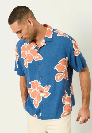 Mann trägt ein blaues kurzärmeliges Hemd mit großen orange-weißen Blumenmustern und schaut nach unten, die Hände in den Taschen.
