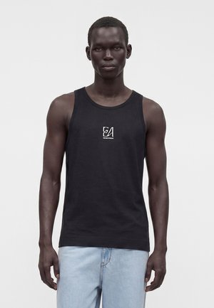 Homme portant un débardeur noir avec un petit logo blanc sur la poitrine et un jean bleu clair, debout devant un fond clair uni.