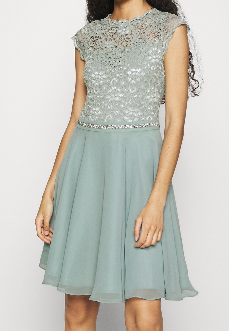 Femme portant une robe bleu sarcelle clair avec un corsage en dentelle, des manches courtes, une ceinture scintillante et une jupe fluide en mousseline, debout devant un fond uni.
