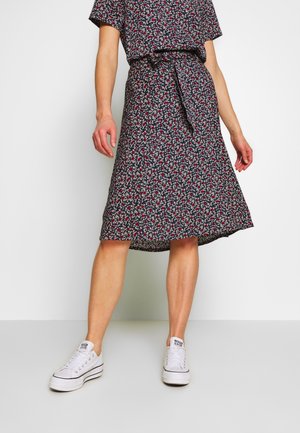 Kvinde iført en navyblå blomsterprint midi nederdel med taljebånd, kombineret med hvide lave sneakers mod en ensfarvet hvid baggrund.