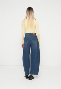 Jeans de denim azul de talle alto con piernas anchas, cinco bolsillos y un parche de marca en la parte trasera. Textura suave, costuras estándar.