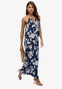 Vestido maxi azul marino con estampado floral en blanco y azul claro, escote en V y tirantes finos. Combinado con sandalias de tacón transparente y un clutch de tejido.