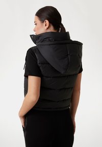 Gilet piumino nero con colletto alto e cappuccio, caratterizzato da un design trapuntato e tasche con zip. Il materiale appare lucido e isolato.