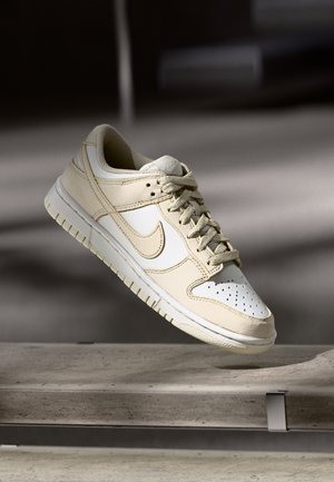 Sneakers Nike beige con tomaia in pelle, puntale perforato e suola ammortizzata. Presentano uno swoosh bianco e dettagli testurizzati.