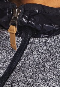 Giacca in pile con un design testurizzato grigio e nero, dotata di un colletto nero lucido e una cerniera con un tirante beige per un facile accesso.
