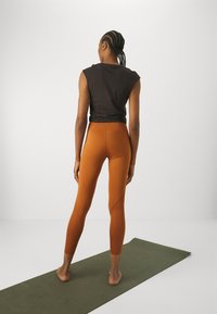 Fekete ujjatlan felső és narancs színű leggings sima textúrával, testhezálló kialakítással és ferde oldalpanelekkel. Meztelen lábbal egy zöld szőnyegen viselve.