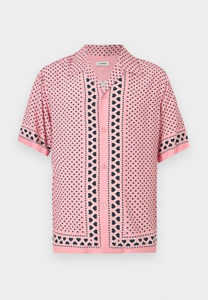 Mand står mod en ensfarvet væg iført en pink mønstret kortærmet skjorte, sorte bukser med vide ben og hvide sneakers.