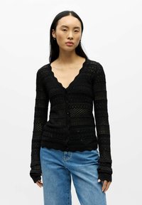 Schwarze Strickjacke mit V-Ausschnitt, strukturiertem Strickmuster und gewellten Kanten. Hat lange Ärmel und einen Knopfverschluss. Kombiniert mit hellblauen Jeans.